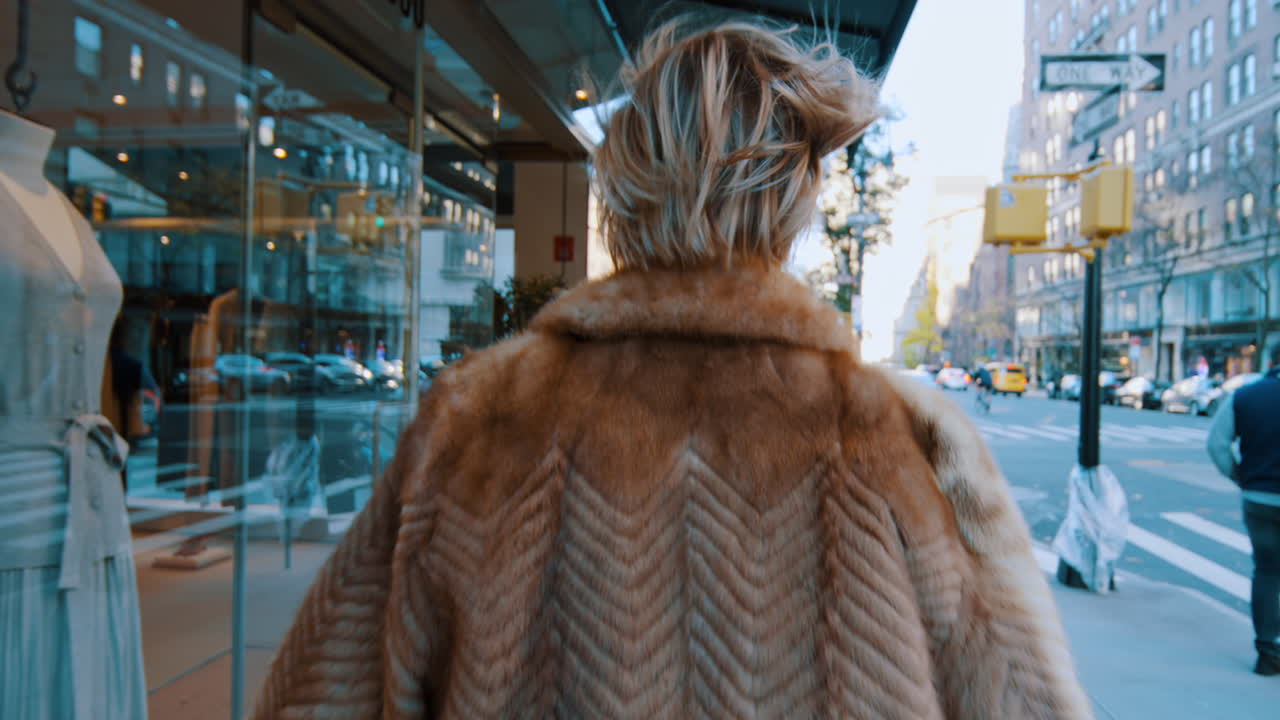 New York, USA, 5 ottobre 2025: Donna elegante con pelliccia che cammina su un marciapiede di Manhattan. Vista dal basso di una donna con pelliccia marrone e occhiali da sole che cammina davanti a una vetrina di vetro in una strada di New York.