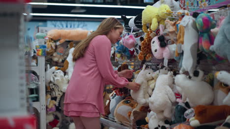 lady in pink dress explores a toy store while holding a toy, she squats to inspect and feel the texture of a plush toy, surrounded by various colorful toys on the shelves, the store is brightly lit