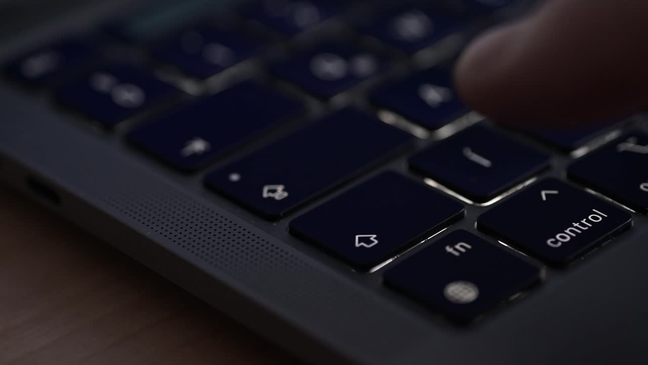 A close-up video of a person's finger pressing the right arrow key on a sleek laptop keyboard. The video highlights the dark keycaps, modern design, and the perforated speaker grille on the device