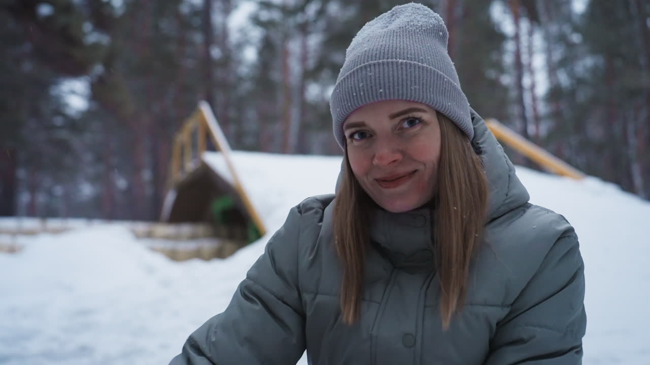 Giovane donna sorride calorosamente mentre si gode una giornata invernale all'aperto, indossando un cappello di lana grigio e un piumino spesso, neve soffice che cade intorno, circondata da una foresta innevata e un ponte di legno in uno sfondo pacifico.