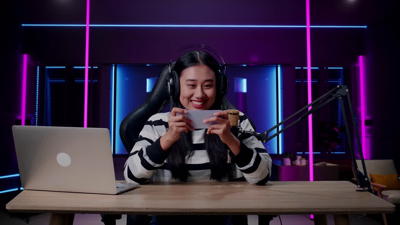 una mujer asiática sonriente con auriculares y computadora usando un teléfono móvil jugando un juego en una habitación iluminada por luces de neón en retro