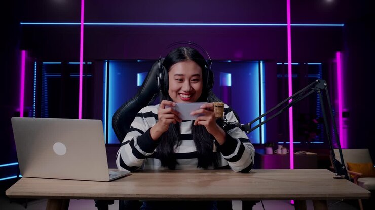 A Smiling Asian Woman Gamer With Headphone And Computer Using Mobile Phone Playing Game In Room Lit By Neon Lights In Retro