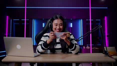 a smiling asian woman gamer with headphone and computer using mobile phone playing game in room lit by neon lights in retro