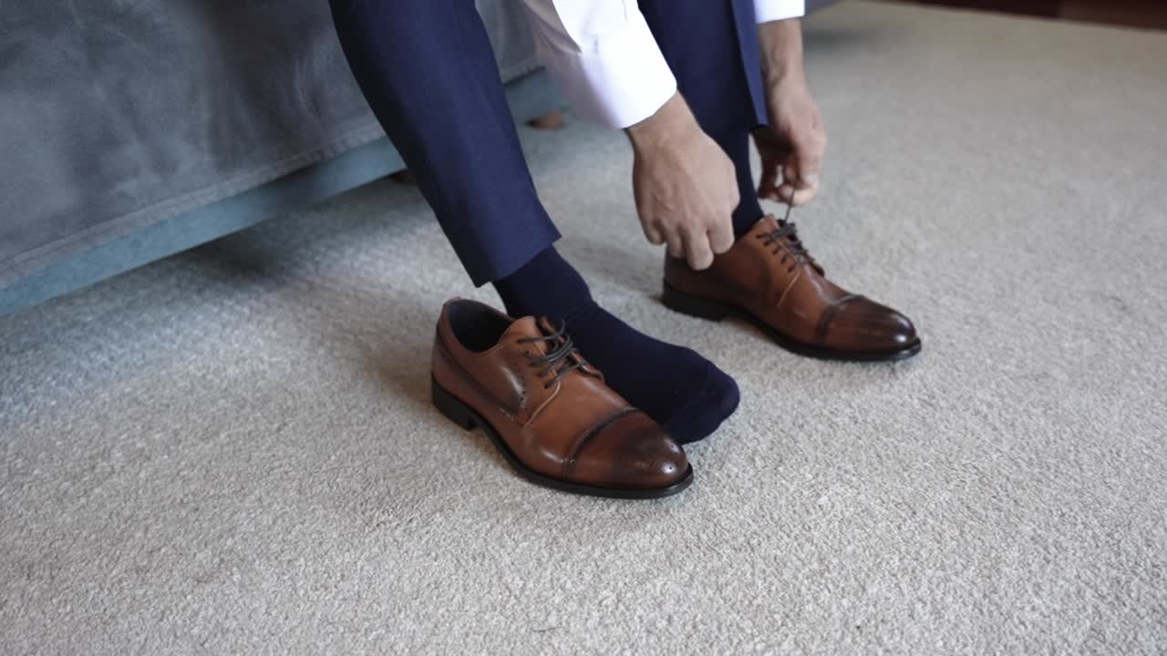Man in navy blue trousers putting on brown leather shoes, seated on a carpeted floor in preparation for a formal event
