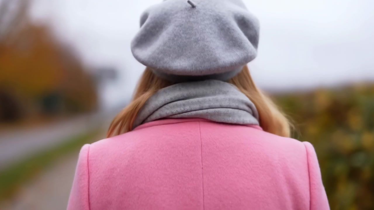 灰色のベレー帽とピンクのコートを着た女性の後ろ姿。ぼやけた道、自然光、穏やかな屋外の雰囲気が漂う柔らかな秋の情景です。