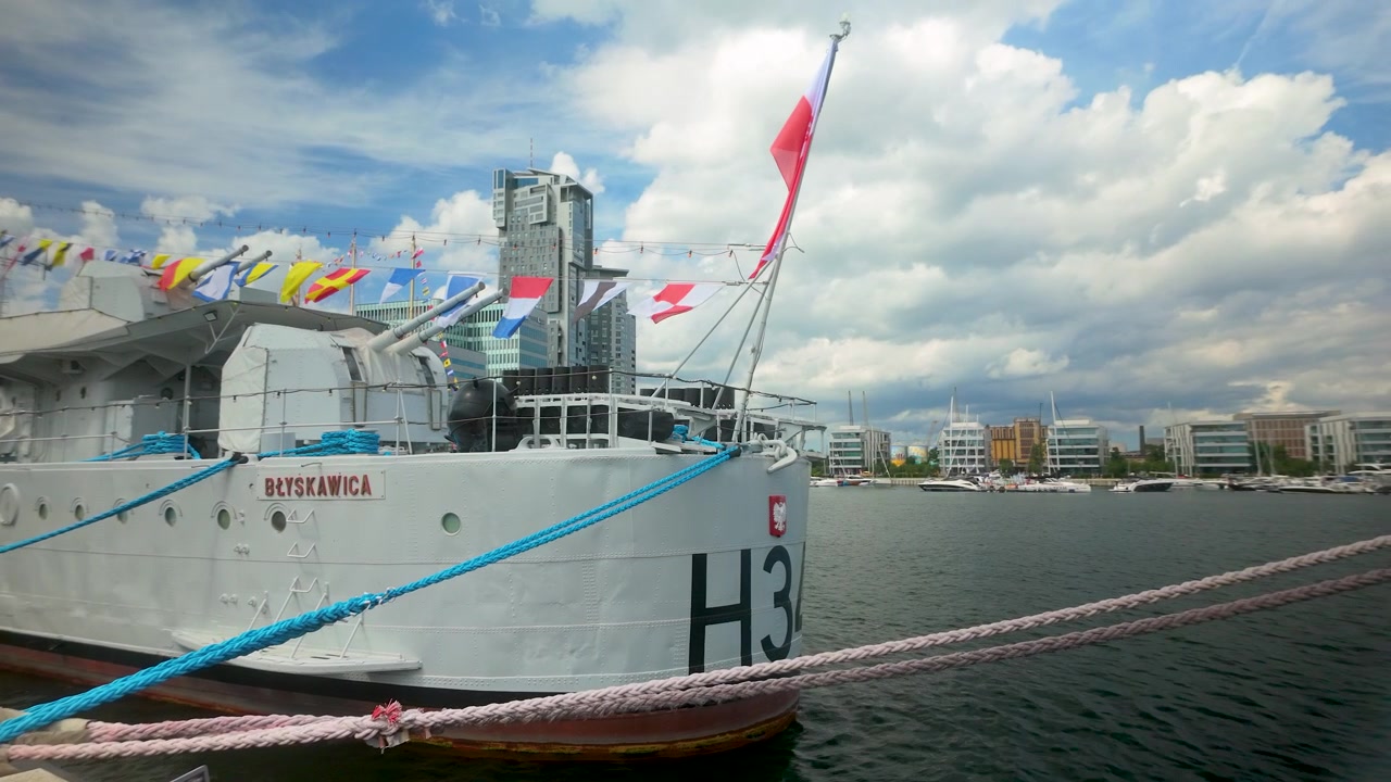 Et vidvinkelbilde av museumsskipet ORP Blyskawica fortøyd ved kaien i Gdynia, med marineflagg og byens silhuett