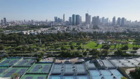 Langsamer-Flug-über-Tennisplätze-Mit-Einem-Malerischen-Stadtbild-Aus-Park-Und-Fluss-In-Der-Nähe-Von-Wohntürmen