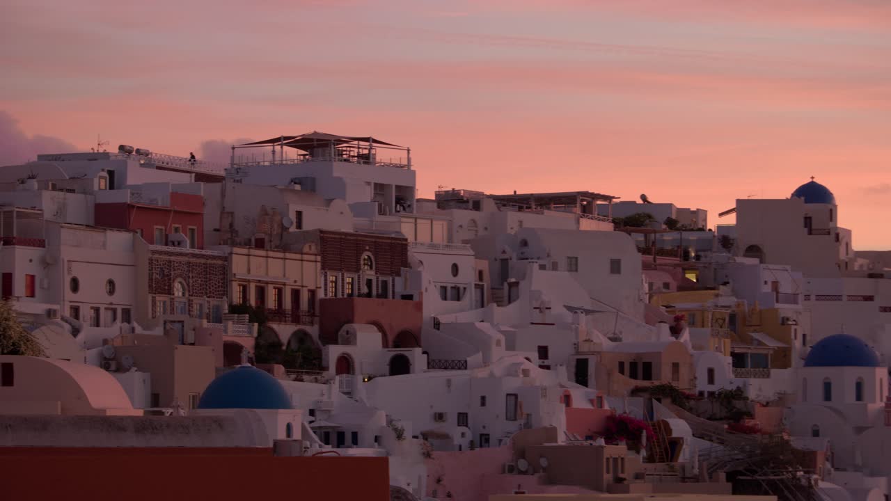 A soft morning glow bathes Santorini’s layered rooftops and colorful domes, capturing the peaceful awakening of this Cycladic village under a pastel sky at sunrise