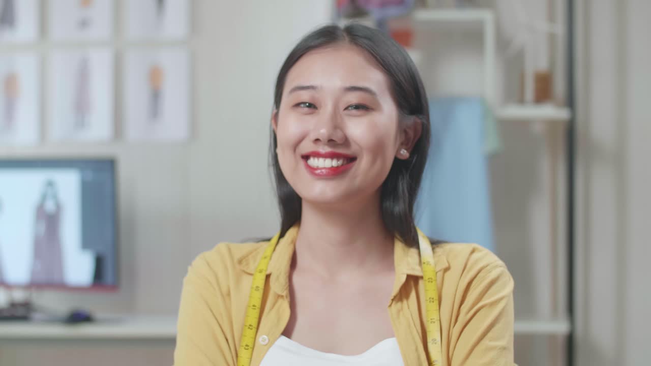 스튜디오에서 옷을 디자인하는 동안 카메라에 미소 짓는 목에 측정 테이프를 가진 아시아 여성 디자이너의 클로즈업