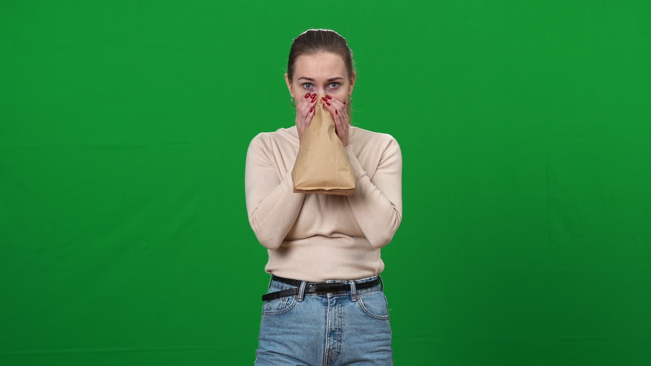 공황상태에 빠진 여성이 종이 봉투에 숨을 들이마시며 녹색 화면에서 카메라를 바라보고 있다.