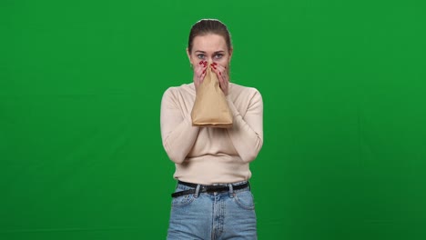 공황상태에 빠진 여성이 종이 봉투에 숨을 들이마시며 녹색 화면에서 카메라를 바라보고 있다.