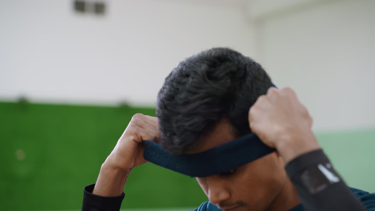Focused athlete with earring lowers head while adjusting blue headband under soft fluorescent indoor light, preparing for sports activity, green blurred background