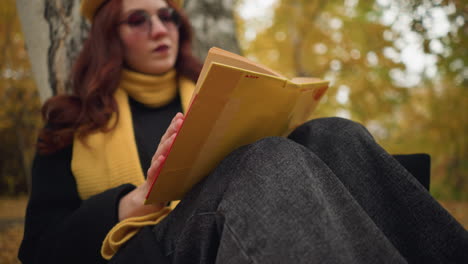 close-up of lady wearing sunglasses and warm scarf, thoughtfully reading yellow-covered book, her hand gently rests on pages while background features softly blurred trees