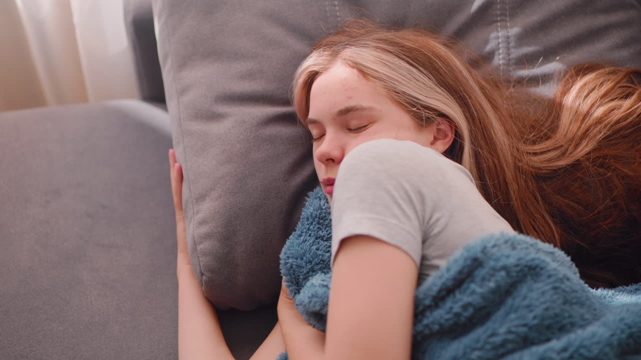 Vista dall'alto di una donna rilassata che dorme con i capelli lunghi appoggiati sul divano mentre abbraccia delicatamente una morbida coperta sotto una calda luce interna, creando una scena calma e confortevole di pura serenità e assoluto relax