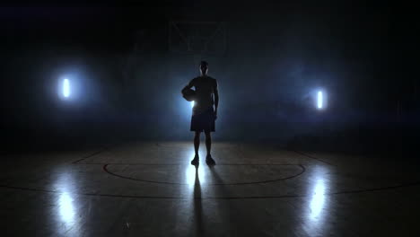 the basketball player stands on a dark playground and holds the ball in his hands and looks into the camera in the dark with a backlit in slow motion and around smoke