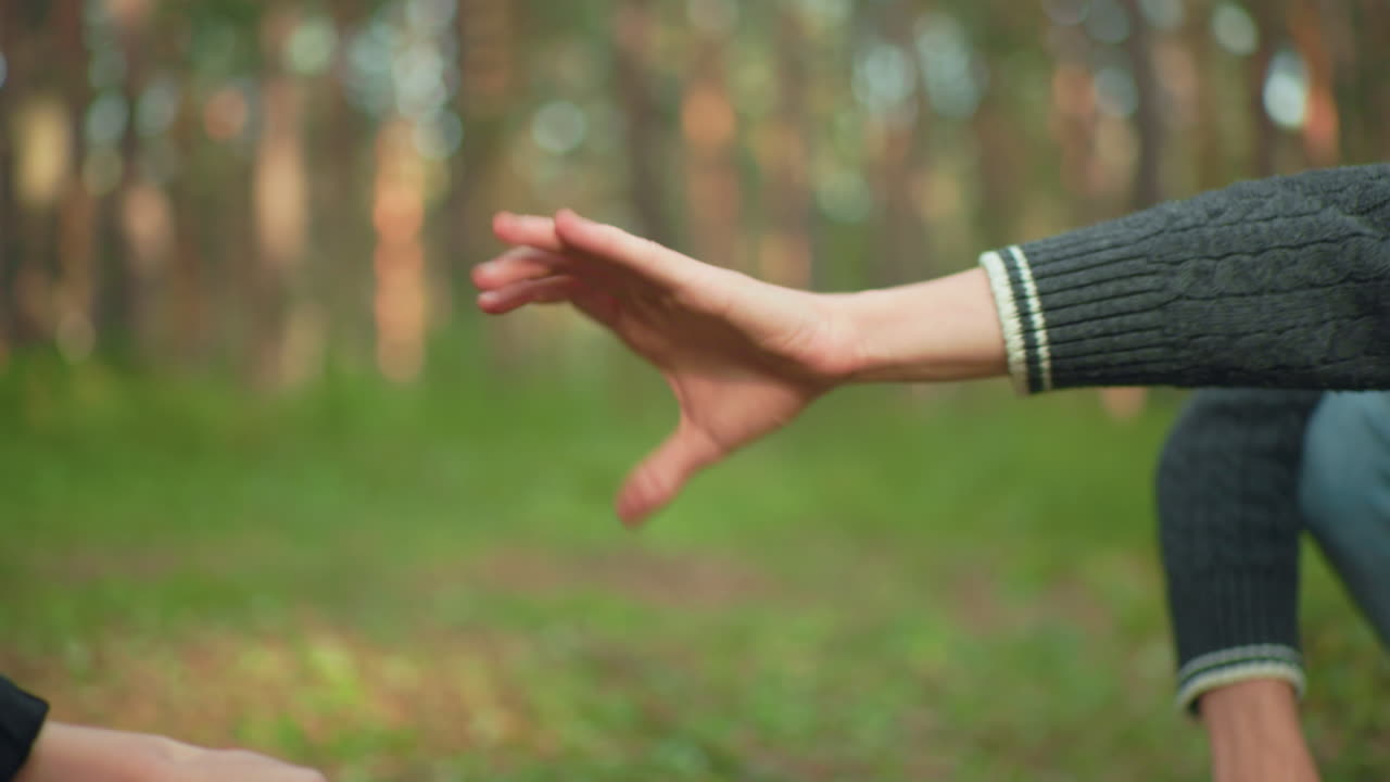 Close up of playful moment between two campers squatting in forest, one reaching to collect tent pole while other holds it back teasingly before eventually handing it over during setup process