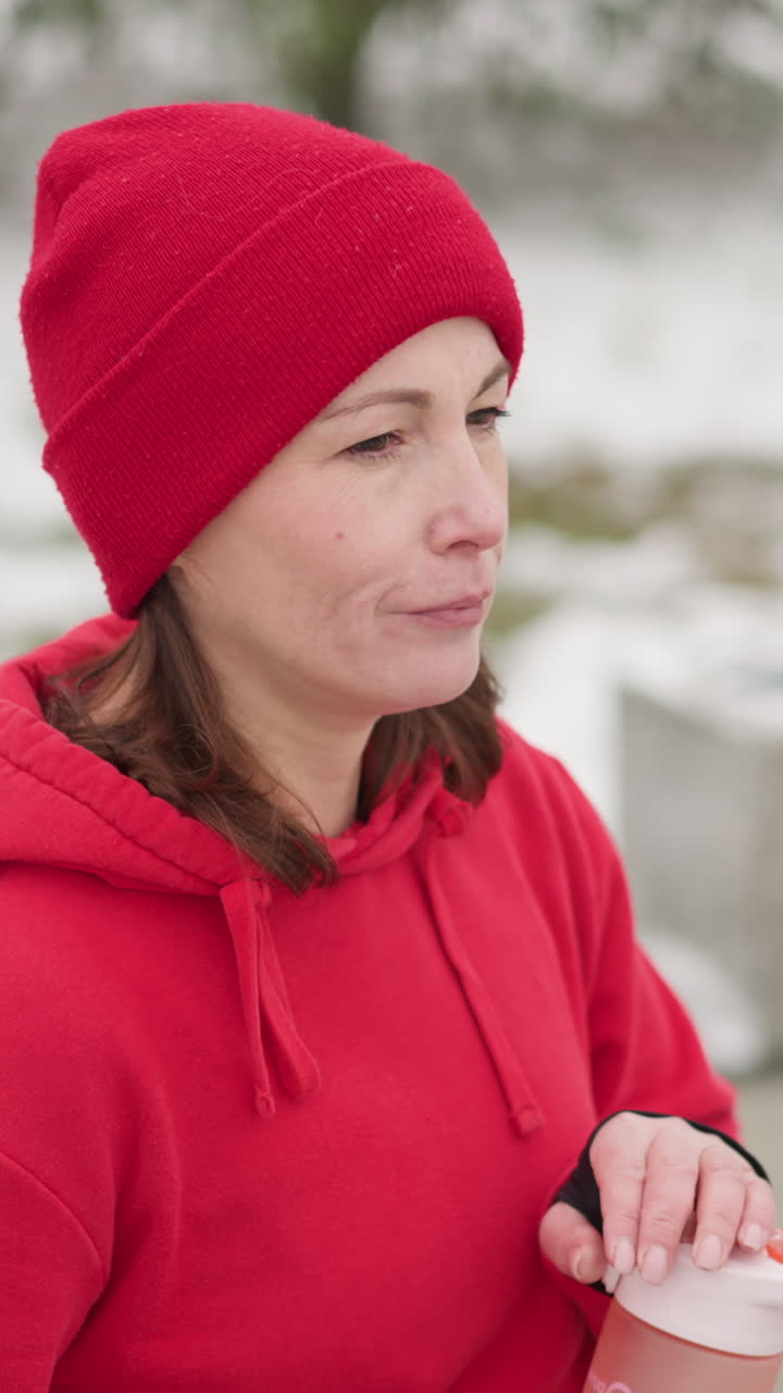 focused woman in red hoodie and beanie closes pink water bottle outdoors, set against snowy background with greenery and blurred concrete bench in serene winter setting