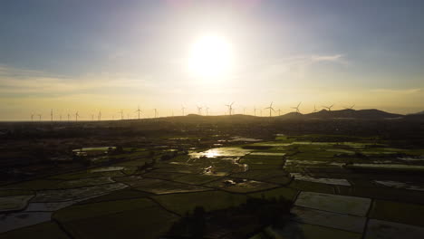 Vista-Aérea-De-La-Puesta-De-Sol-Sobre-Campos-De-Arroz-Agrícolas-En-El-Barrio-De-Lien-Huong-Con-Turbina-De-Molino-De-Viento-Alineada-En-El-Horizonte-Hasta-Las-Colinas-Paisaje-Rural-Vietnamita