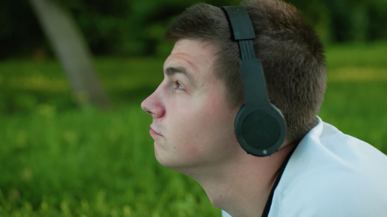 Close up of man lying on grassy field wearing headphones, head slightly lifted, nodding to music while surrounded by lush green grass, embracing tranquility and connection with nature