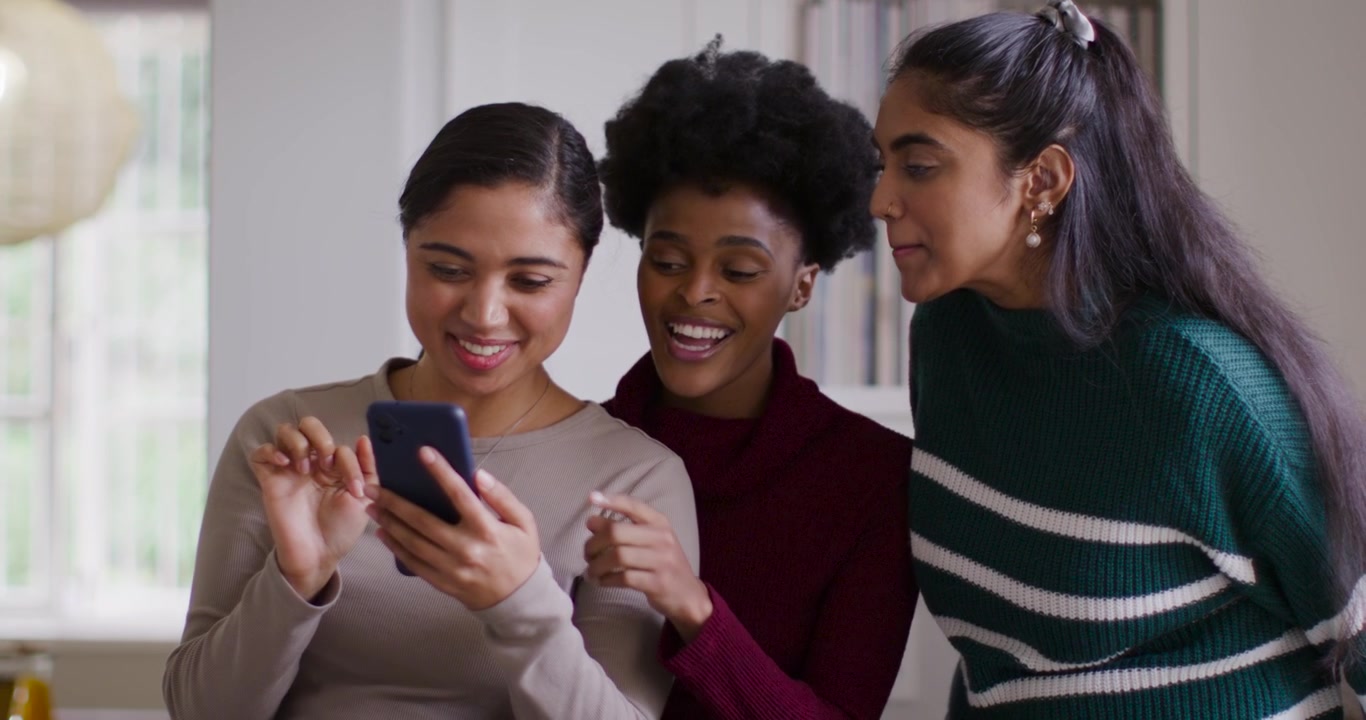 Diverse female friends at home leaning as left woman holding smartphone tapping and sharing content