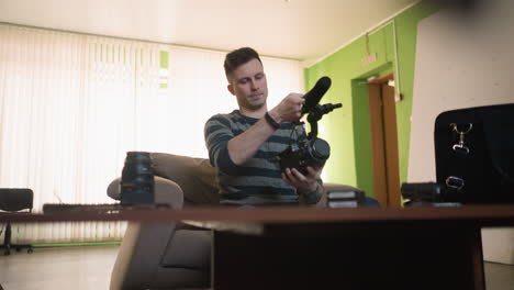 man reaches for camera gear in home studio, organizing equipment in black bag. wooden table filled with camera lenses, microphone, and other tools. cozy green workspace with shelves and clock