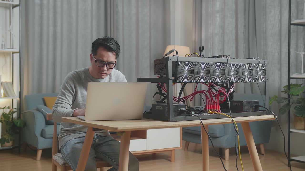 안경을 쓴 진지한 아시아인 남자가 암호화폐를 채굴하기 위해 노트북과 채굴 기계를 사용합니다.