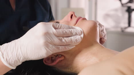Close up view of treatment provider using cotton pads to swab patient jawline to forehead during facial therapy session in bright clinic room with soft ambient lighting and calm atmosphere
