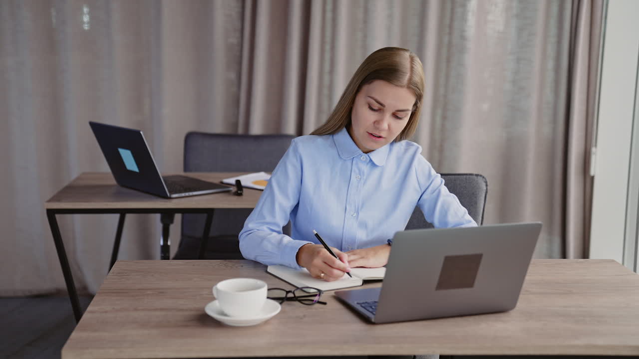Young lady busy with her work in the office. Woman works on laptop and takes notes into her paper notebook.