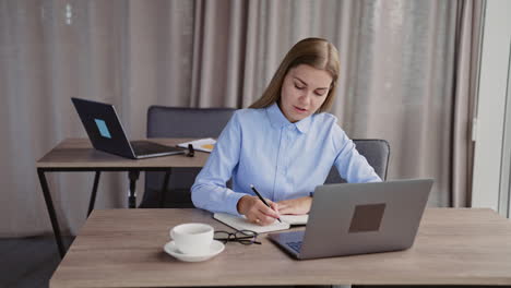 young lady busy with her work in the office. woman works on laptop and takes notes into her paper notebook.
