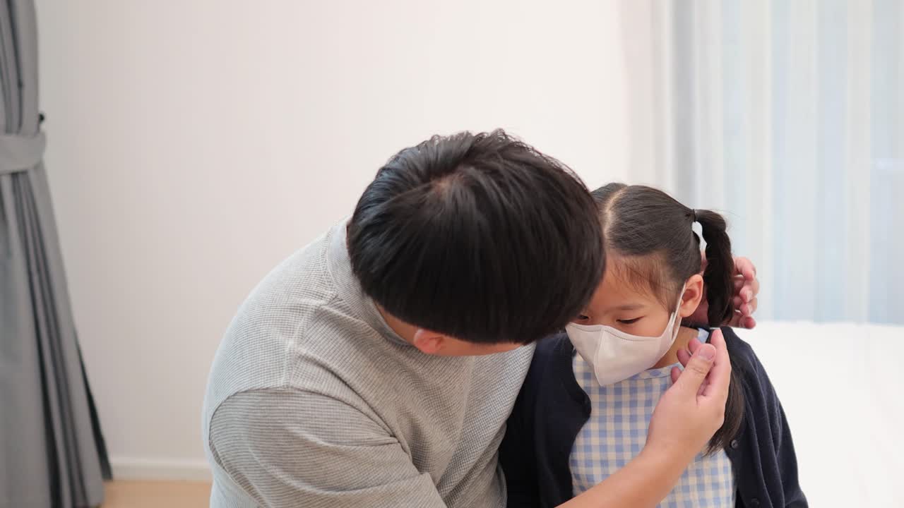 Father assists ill young daughter with wearing a face mask in softly lit bedroom setting