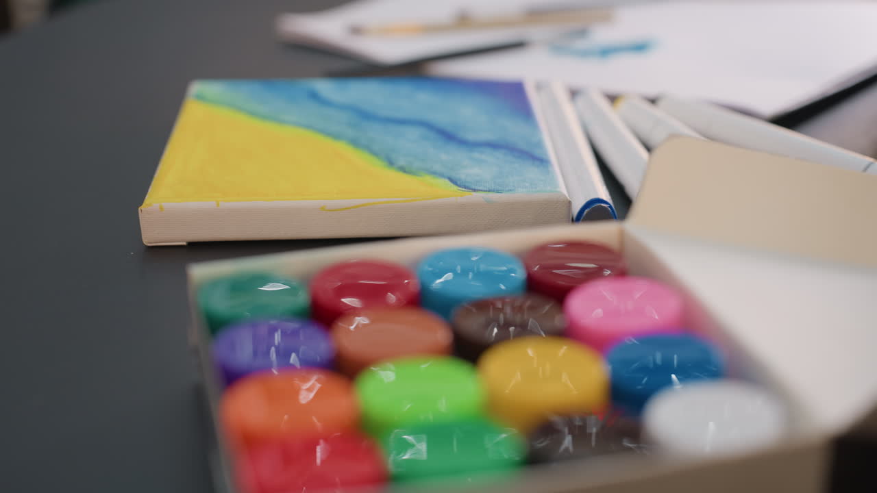 레이어드된 색상과 미술 용품이 있는 작은 텍스처 캔버스의 클로즈업 뷰, 파란색과 노란색 워시가 표현된 미니어처 텍스처 캔버스와 흐릿한 페인팅 도구의 근접 뷰