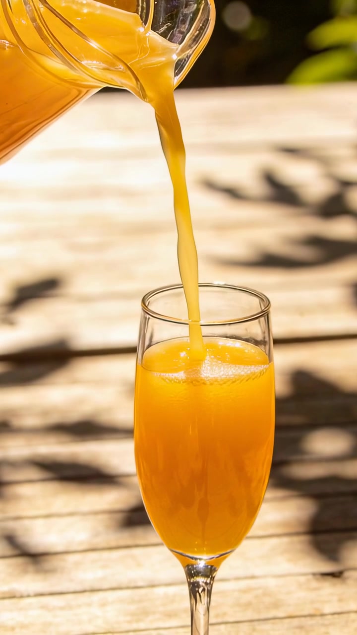Video verticale: Brocca che versa succo d'arancia in un bicchiere a stelo alto su un patio, per il servizio.