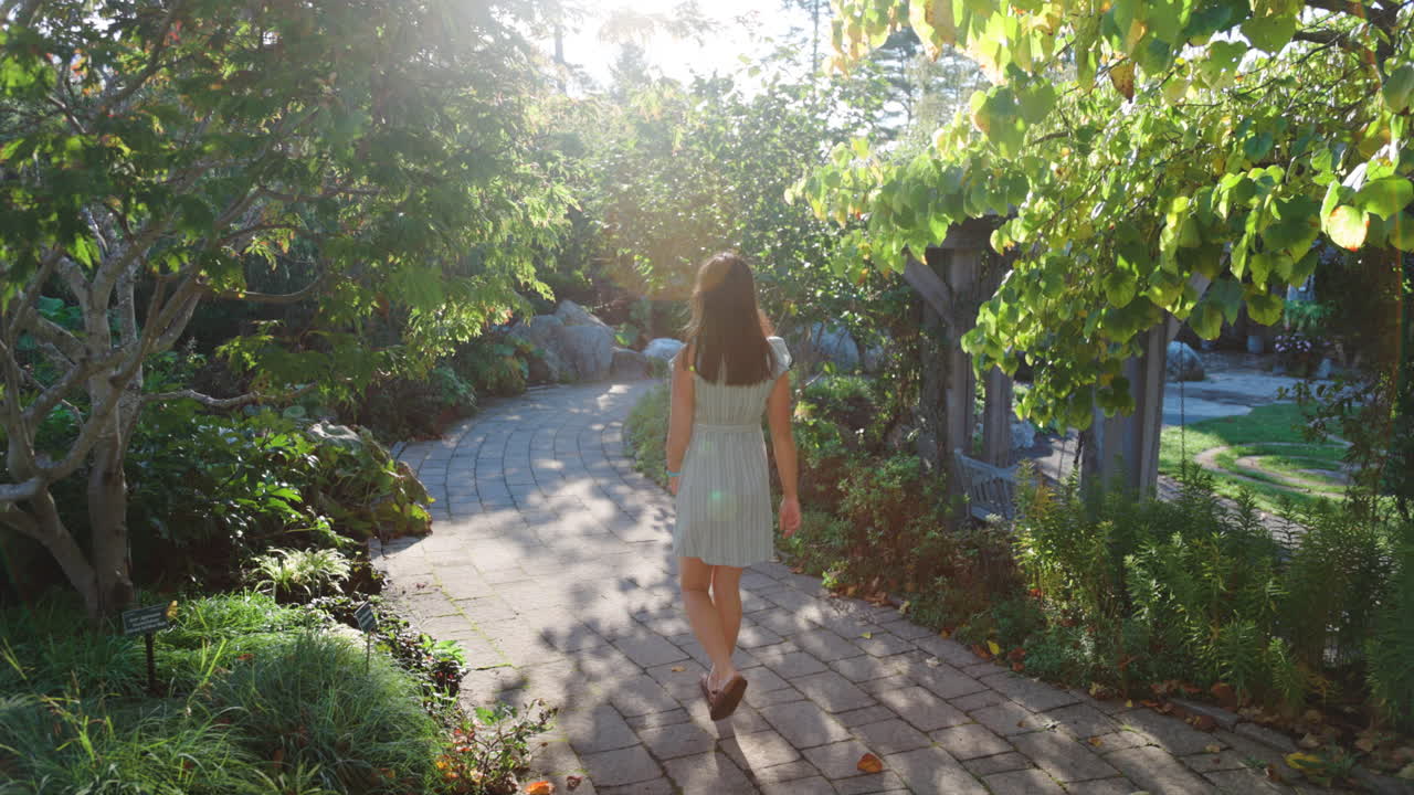 긴 검은 머리와 여름 드레스를 입은 여성이 화창한 날 활기찬 공원에서 걸어가고 있습니다.