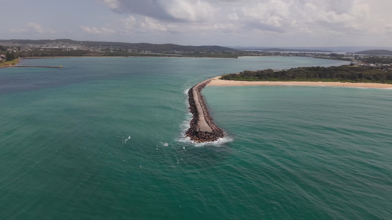 Drone orbits over breakwall meeting blue green sea off sunny Blacksmiths Beach coast, NSW Australia