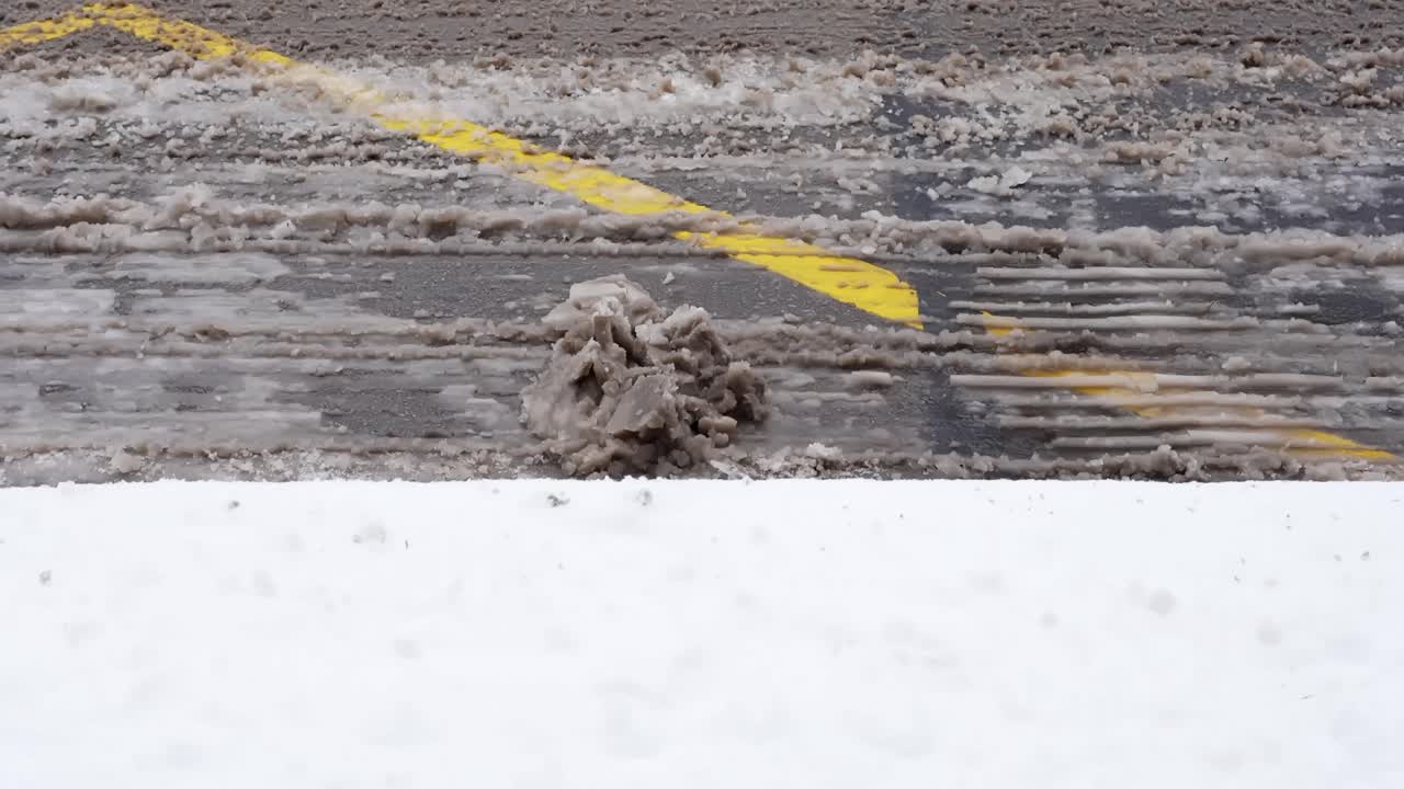 Panoramica dal basso verso l'alto e primo piano di una strada asfaltata fangosa con chiazze di neve sporca, acqua di disgelo e una segnaletica orizzontale gialla, che mostra le difficili condizioni stradali invernali