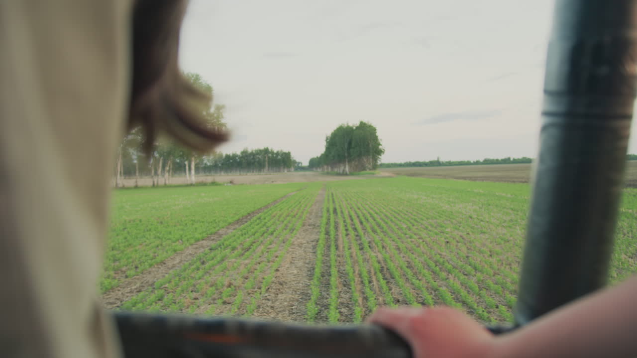 close up from moving hot air balloon showing expansive green farmland below with hand resting on basket edge and rows of crops stretching into distance under soft sky