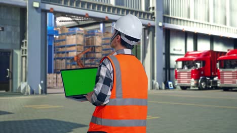 back view of asian male engineer with safety helmet working on a green screen laptop and looking around while standing , outside of logistics distributions warehouse