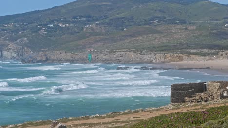 SLOW-MO-wing-surfer-on-azure-waves-with-mountains-in-the-background,-Cape-Roca,-Portugal