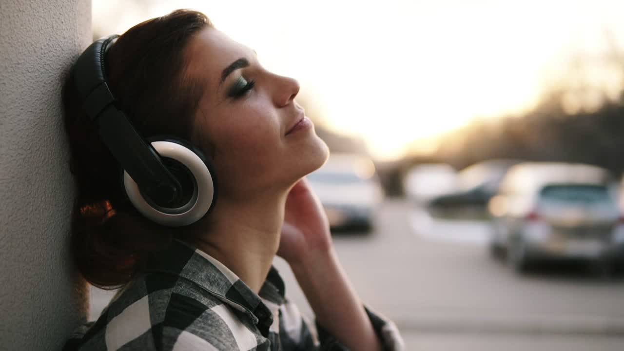 fondo borroso brillante al aire libre. vista lateral de una hermosa chica poniendo auriculares y relajándose. escuchar la música. apoyarse en la pared. sonriendo