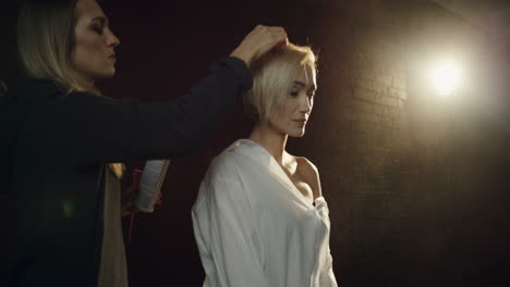 hairdresser doing hair of the blond female model during her photoshoot in the dark studio with a single spotlight