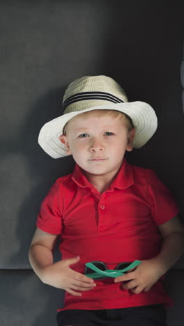 little boy tourist in hat takes off sunglasses looking in camera with smile. cute toddler in red t-shirt lies on sofa near suitcase upper closeup