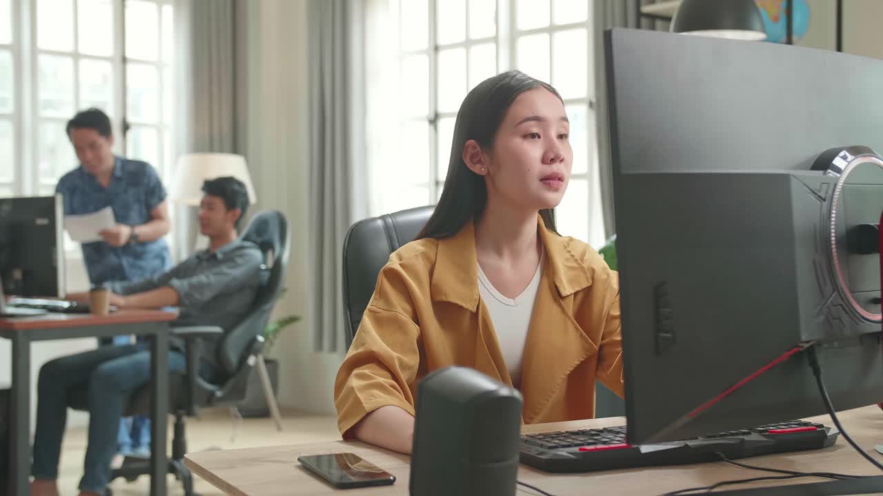 Asian Female Drinking Coffee While Works On Her Personal Computer With Big Display In Office