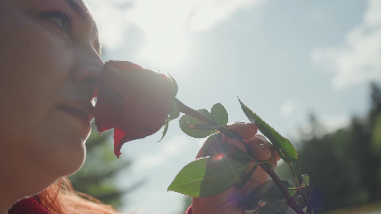 Primo piano di una donna che tiene in mano e annusa una rosa rossa all'aperto, con la luce del sole che illumina il suo profilo mentre avvicina il fiore al naso. Lo sfondo presenta vegetazione sfocata sotto un cielo blu.
