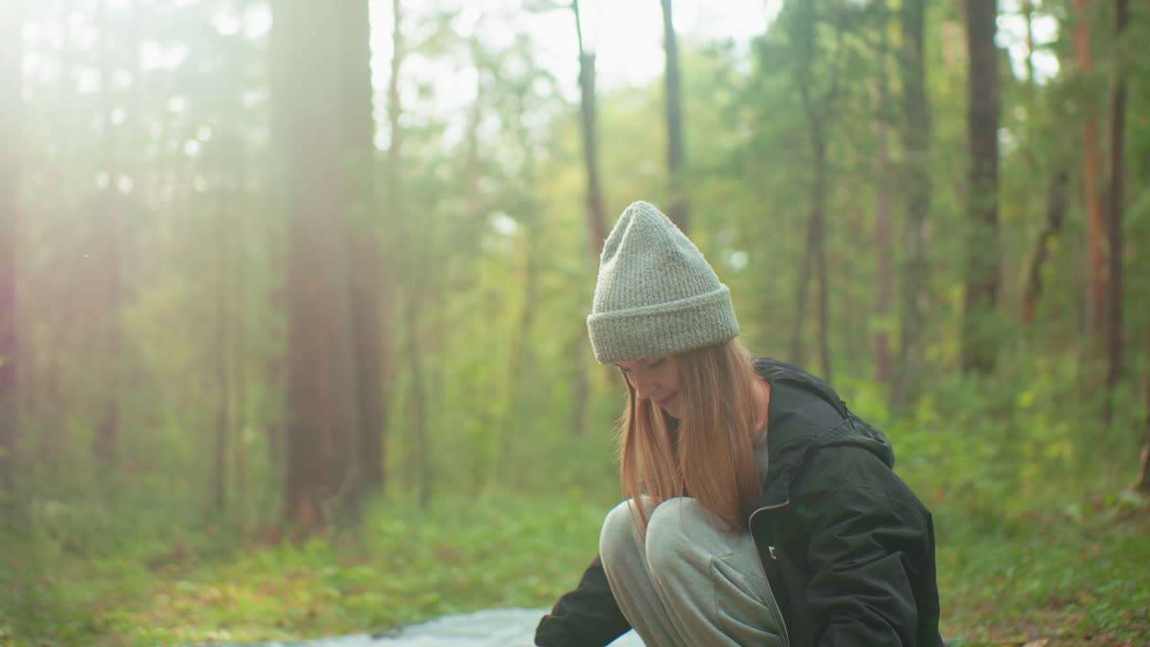 Young woman wearing beanie and black jacket crouches on forest ground while preparing tent pole for camping, soft sunlight glows through trees creating peaceful outdoor atmosphere