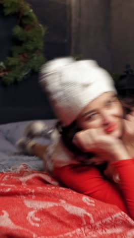 emotional girl at christmas. beautiful young woman in knitted cap and scarf laying on bed on christmas presents and fir branches background. vertical video