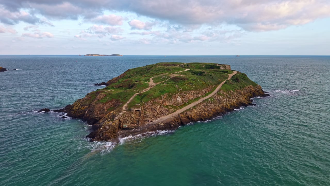 Grand Bé island off Saint-Malo, Brittany, France, showing rocky cliffs, grassy top and walking path, with Fort du Petit Bé in background, drone orbit shot.