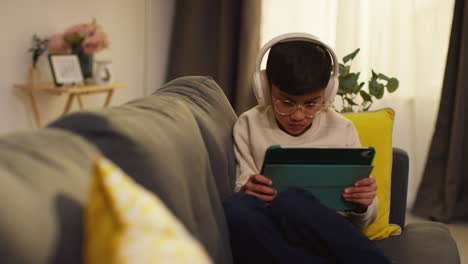 young boy sitting on sofa at home wearing wireless headphones playing games or streaming onto digital tablet 1