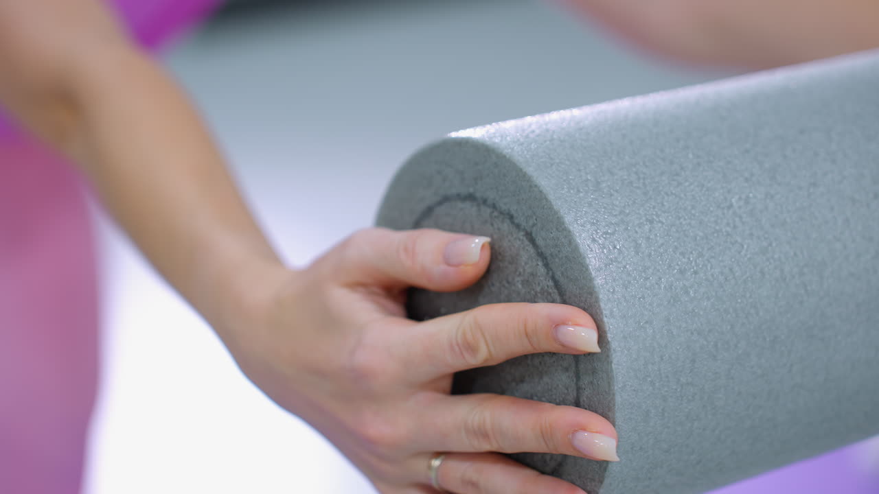 primer plano de una mano de mujer sosteniendo una espuma de rodillo, con un anillo en su dedo, mientras empuja hacia adelante, con una vista parcial de otra persona en el fondo en un entorno de gimnasio