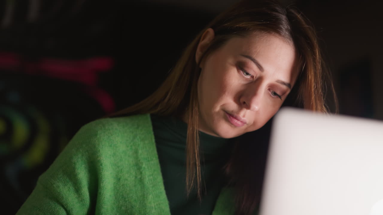 Una donna con un maglione verde che lavora al laptop con un'espressione concentrata e pensierosa. Ripresa con una telecamera a mano, enfatizzando la concentrazione e uno spazio di lavoro digitale moderno.