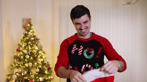 Happy-young-man-with-a-ho-ho-ho-sweater-grabs-a-Santa-hat-in-front-of-a-Christmas-tree,-throwing-a-Christmas-hat-in-slow-motion